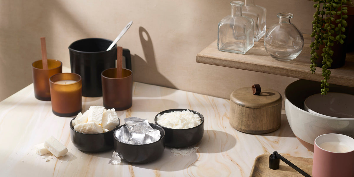 A cozy candle-making setup with amber jars, bowls of wax, and glass containers on a warm marble surface. Natural light highlights the tools, wooden accents, and greenery for an inviting, handcrafted feel.