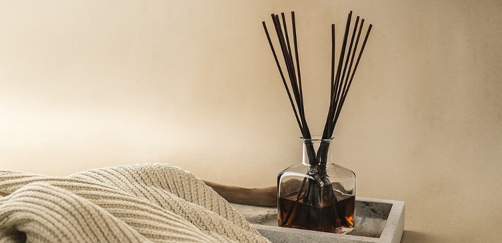 A glass reed diffuser sits beside a soft knit blanket, creating a cozy and inviting home fragrance scene.