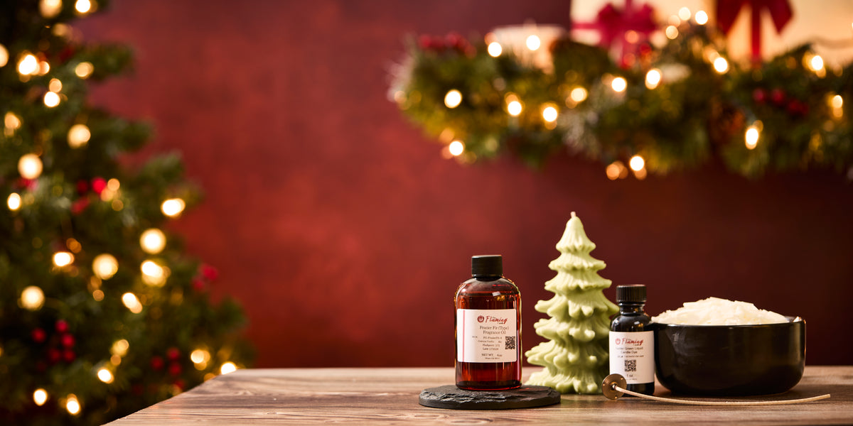 A holiday-themed product scene featuring The Flaming Candle fragrance oils and candle-making supplies arranged on a wooden surface. A red-tinted bottle of fragrance oil sits on a black coaster next to a small green Christmas-tree-shaped candle. A black bowl filled with white wax and a smaller bottle of fragrance oil sit nearby with a wick tool resting in front. In the background, warm Christmas lights, garland, and a decorated tree create a festive, cozy glow.