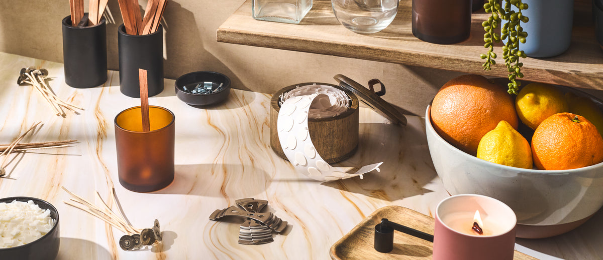 A candle-making workspace with wicks, metal wick holders, soy wax flakes, adhesive tabs, and amber glass jars arranged on a marble surface beside a bowl of citrus fruits and a lit candle in a pink vessel.