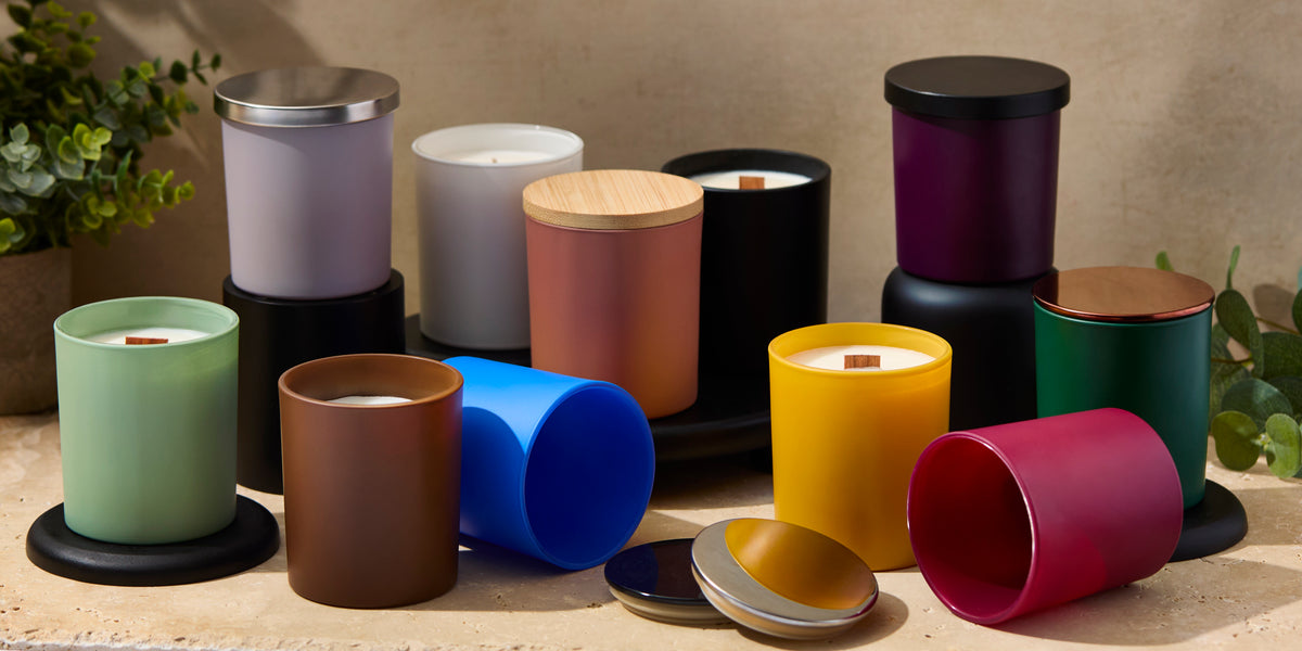 Assorted colorful candle jars displayed upright and on their sides against a warm neutral backdrop, with greenery and wooden-wick candles throughout.